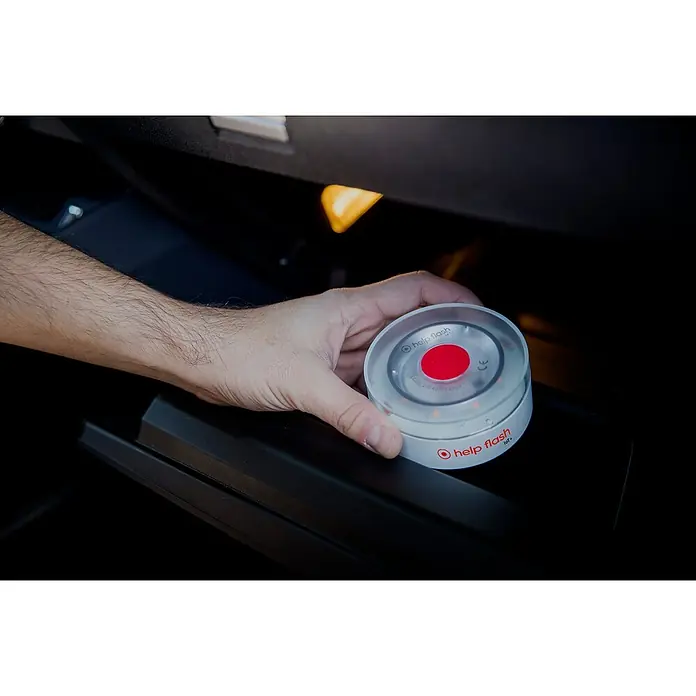 Luz de emergencia Help Flash IoT+ - Apto para: Avería en el vehículo - Una mano coloca una luz de emergencia redonda Help Flash IoT con un botón rojo y luces LED en un compartimento oscuro del coche.