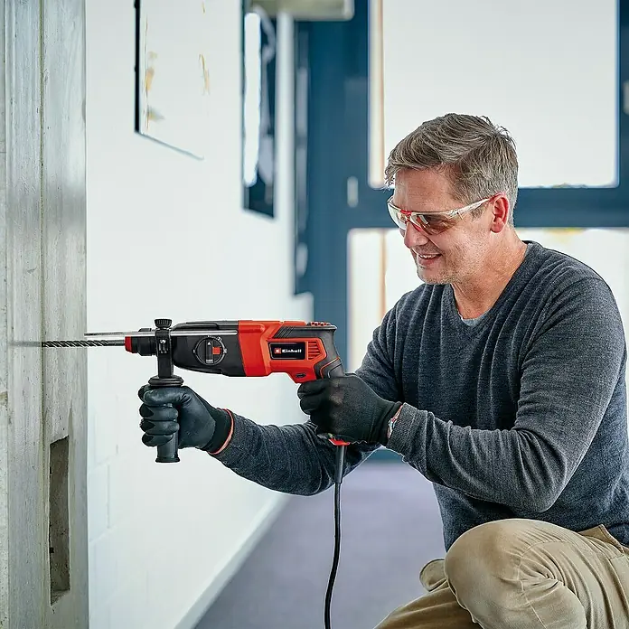 Einhell Kombihammer TC-RH 620 4F Kit - 620 W, 2,2 J, SDS-Plus-Aufnahme - Ein Mann bohrt mit einer Einhell Schlagbohrmaschine ein Loch in eine Wand, wobei er eine Schutzbrille und Handschuhe trägt.