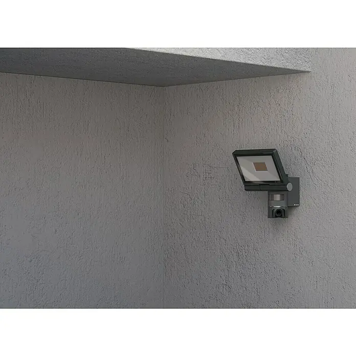 Steinel LED-Strahler CAM 2 SC - 21 W, 2.310 lm, 3.000 K, IP44 - Nahaufnahme einer modernen, grauen Außenleuchte mit integrierter Überwachungskamera an einer grauen Wand.