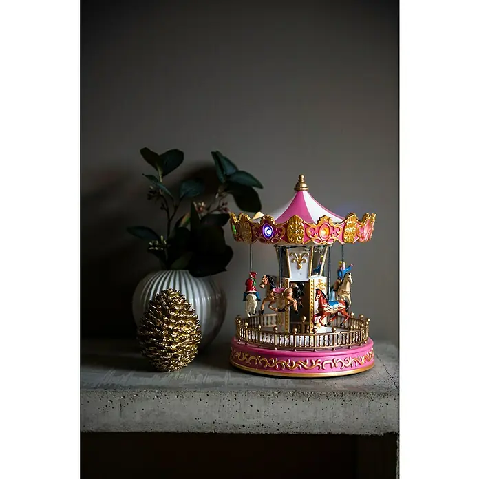 Konstsmide LED-Szenerie Weihnachtskarusell - L x B x H: 18,2 x 18,2 x 23,5 cm, Rosa, Batteriebetrieben, LED - Die Aufnahme zeigt ein detailliertes Karussell mit rosa und weißen Akzenten, umgeben von dekorativen Elementen auf einem grauen Tisch.