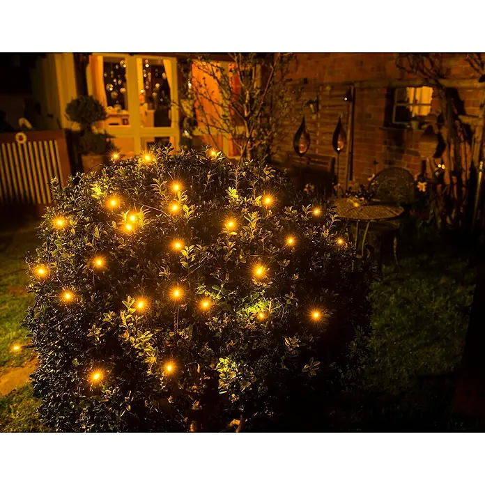 Konstsmide LED-Lichtnetz - Außen, Lichtfarbe: Bernstein, Netzbetrieben - Ein üppiger Busch, geschmückt mit warm leuchtenden Lichterketten, erhellt stimmungsvoll den Garten und schafft eine einladende Atmosphäre.