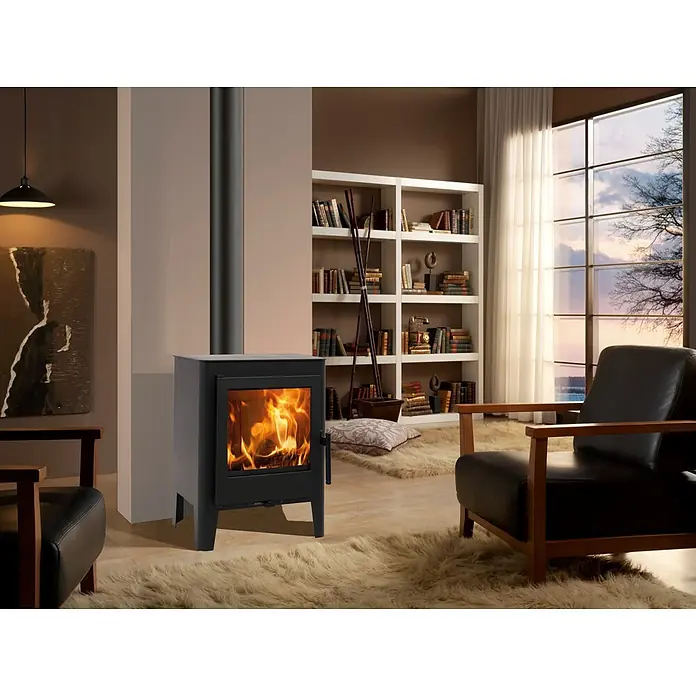 Panadero Estufa de leña Majster - 5,9 kW, Volumen de calefacción: 180 m³ - Una imagen detallada muestra una estufa de leña en la habitación, que irradia calidez y confort, complementada con muebles y libros.