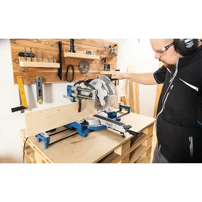 Scheppach Kapp- & Gehrungssäge HM305SL - 2.000 W, Durchmesser Sägeblatt: 305 mm, Leerlaufdrehzahl: 5.000 U/min - Ein Mann bedient eine Scheppach Kapp- und Gehrungssäge auf einer Holzpalette, während er Sicherheitsausrüstung trägt und Holz bearbeitet.