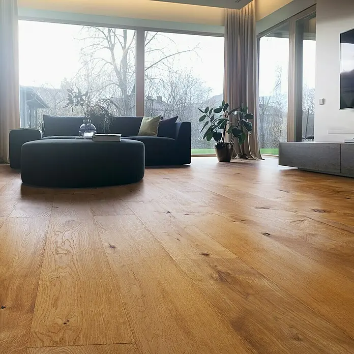 Parkettmuster Eiche Fabia - 300 x 190 x 14 mm, Landhausdiele - Einladendes Wohnzimmer mit Holzboden, grauem Sofa und Ottomane, akzentuiert durch eine Zimmerpflanze und große Fenster für natürliche Beleuchtung.