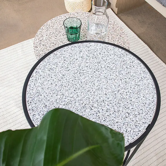 D-c-fix Porrinho-Effektfolie - 200 x 45 cm, Grau-Blau, Selbstklebend - Nahaufnahme von zwei runden Tischen mit Terrazzo-Oberfläche und Getränken auf einem strukturierten Teppich.