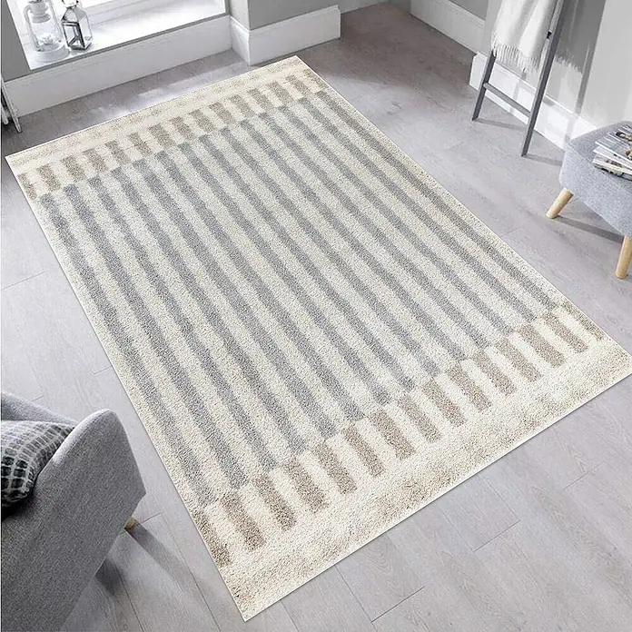 Alfombra de pelo largo Rayas - Natural, 230 x 160 cm, 100% Polipropileno - Una alfombra rectangular con un patrón de rayas en tonos grises y beige se encuentra sobre un suelo de madera clara. La alfombra tiene un borde en los extremos.