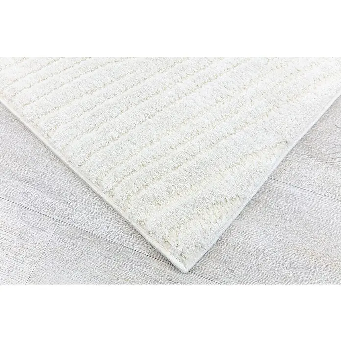 Alfombra de pelo largo Relieve - Crudo, 110 x 60 cm, 80% polipropileno / 20% poliéster - Primer plano de una alfombra blanca con un patrón de rayas sobre un piso de madera clara.