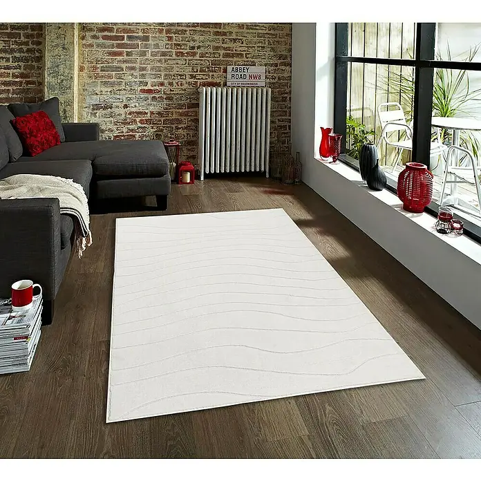 Alfombra de pelo corto Stelvio Olas - Crudo, 200 x 140 cm, 100% Poliéster - Una alfombra beige claro con un patrón de ondas se encuentra en una sala de estar moderna con un sofá gris y pared de ladrillo.