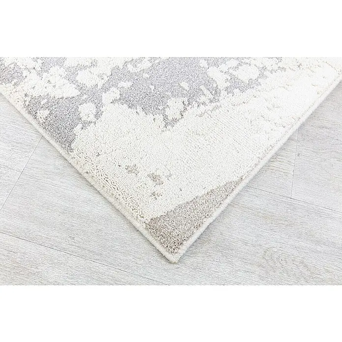 Alfombra de pelo corto Brochazos - Natural, 110 x 60 cm, 100% Poliéster - Primer plano de una alfombra moderna con un patrón abstracto en tonos de gris y blanco, colocada sobre un suelo de madera clara.