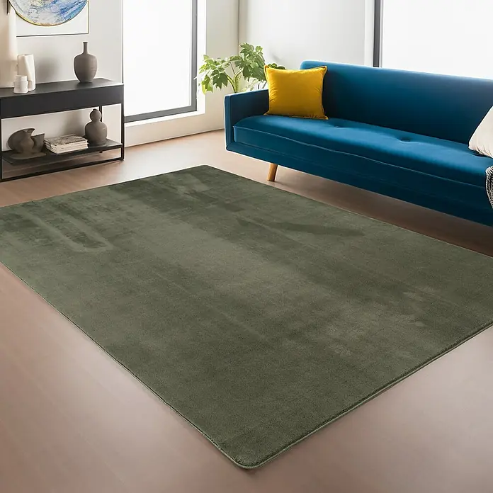 Kurzflorteppich Joy - Grün, 280 x 200 cm, 100 % Polyester - Ein olivgrüner Teppich liegt in einem Wohnzimmer mit einem blauen Sofa und einem schwarzen Regal. Der Teppich verleiht dem Raum Wärme.