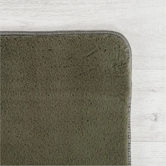Kurzflorteppich Joy - Grün, 280 x 200 cm, 100 % Polyester - Nahaufnahme eines grünen Teppichs mit grauem Rand auf einem hellen Holzboden, der die Textur und Farbe des Teppichs hervorhebt.