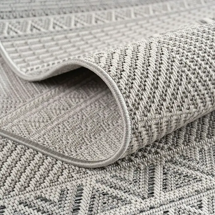 Outdoor-Teppich Desert 1302 - Grau, 290 x 200 cm, 100 % Polypropylen - Detailansicht eines hellgrauen Teppichs mit geometrischem Muster und gerolltem Rand, der die Textur hervorhebt.