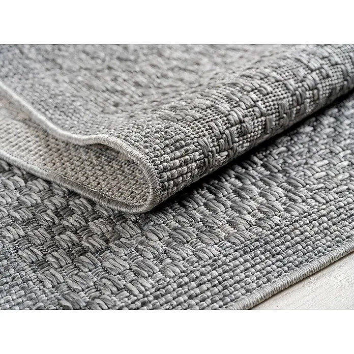 Outdoor-Teppich Ibiza I - Grau, 170 x 120 cm, 100 % Polypropylen - Nahaufnahme eines grauen Outdoor-Teppichs mit gewebter Textur, der teilweise aufgerollt ist und die detaillierte Verarbeitung zeigt.