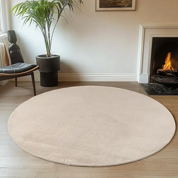 Kurzflorteppich Joy - Creme, Durchmesser: 200 cm, 100 % Polyester - Beiger runder Teppich im Wohnzimmer vor einem Kamin und einem Sessel, der eine warme und einladende Atmosphäre schafft.