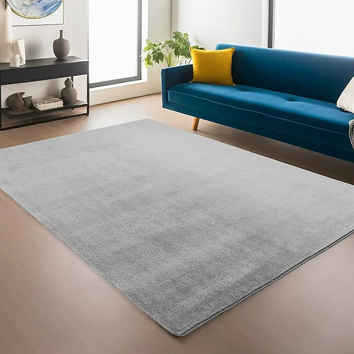 Kurzflorteppich Joy - Silber, 280 x 200 cm, 100 % Polyester - Ein grauer Teppich liegt im Wohnzimmer vor einem blauen Sofa, einem schwarzen Sideboard und großen Fenstern, die viel Licht hereinlassen.