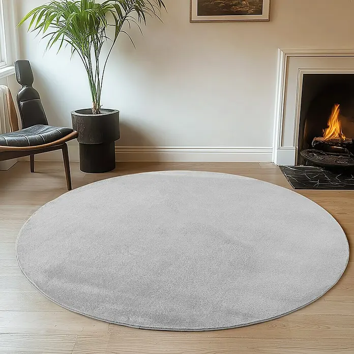 Kurzflorteppich Joy - Silber, Durchmesser: 200 cm, 100 % Polyester - Ein runder, hellgrauer Teppich liegt in einem Wohnzimmer mit Holzboden, Kamin und Zimmerpflanze.