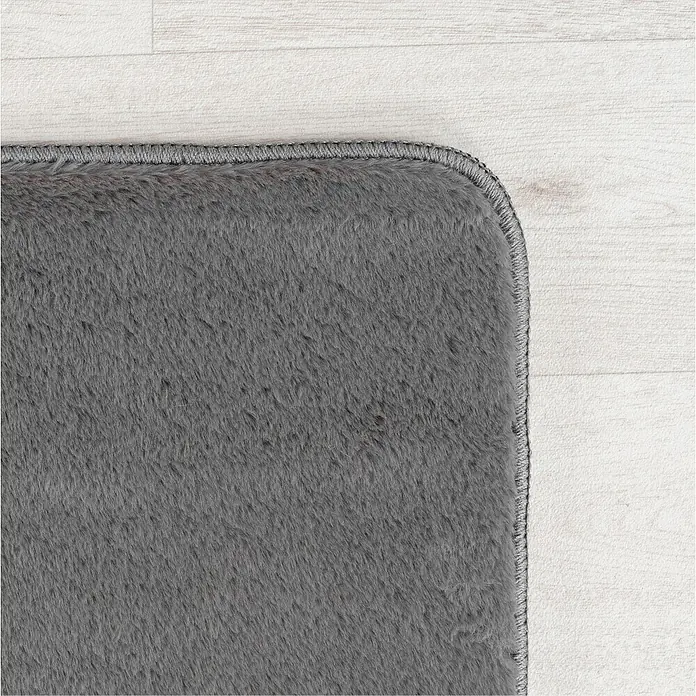 Kurzflorteppich Joy - Grau, 220 x 160 cm, 100 % Polyester - Detailaufnahme eines grauen Teppichs mit abgerundeter Ecke, der auf einem hellen Holzboden liegt. Die Textur des Teppichs ist weich und flauschig.