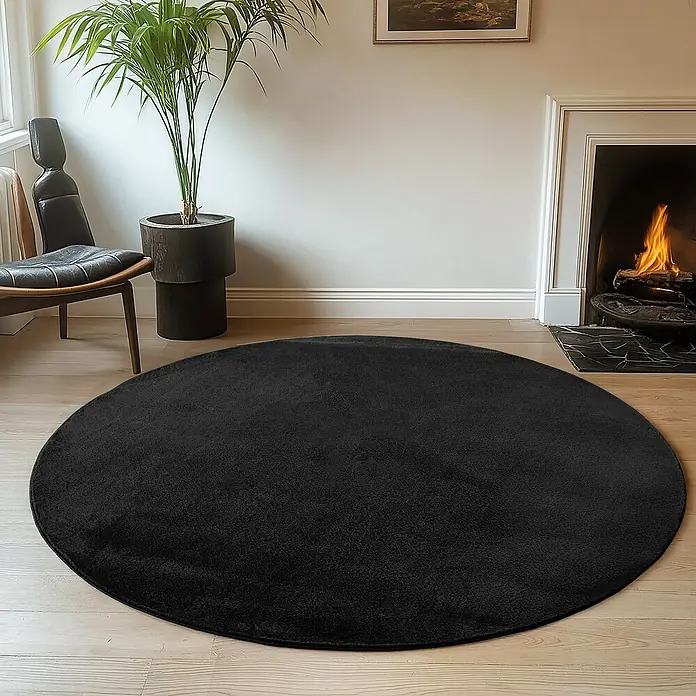 Kurzflorteppich Joy - Schwarz, Durchmesser: 80 cm, 100 % Polyester - Ein schwarzer, runder Teppich liegt in einem Wohnzimmer mit Kamin und Zimmerpflanze.