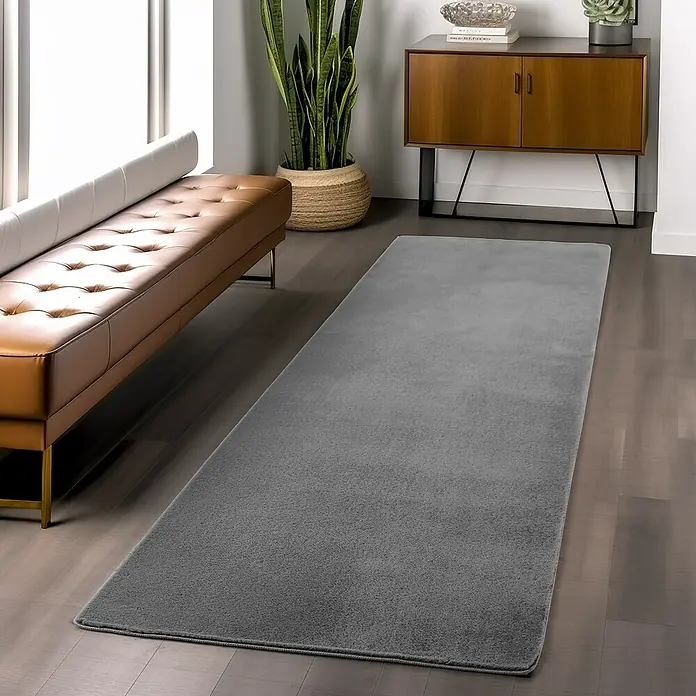 Kurzflorteppich Joy - Grau, 100 x 60 cm, 100 % Polyester - Ein grauer Teppichläufer liegt auf einem dunklen Holzboden in einem modern eingerichteten Raum, der eine elegante Atmosphäre schafft.