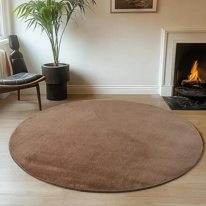 Kurzflorteppich Joy - Mokka, Durchmesser: 200 cm, 100 % Polyester - Ein runder, brauner Teppich liegt in einem Wohnzimmer mit Kamin und Zimmerpflanze.