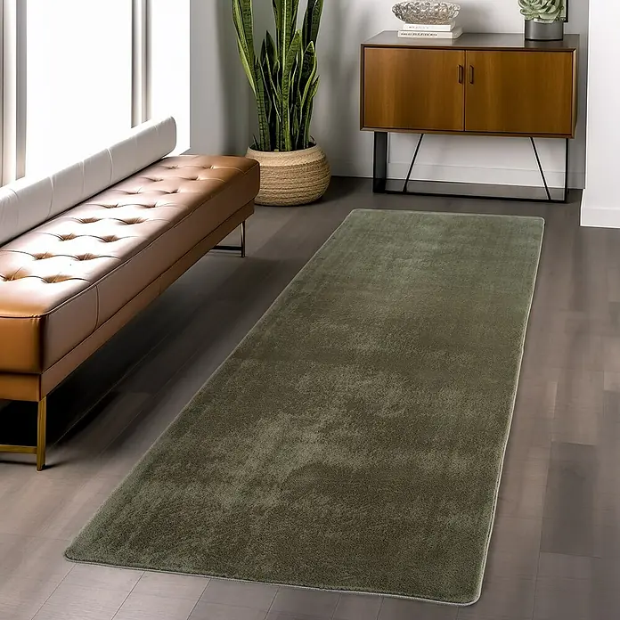 Kurzflorteppich Joy - Grün, 100 x 60 cm, 100 % Polyester - Ein rechteckiger, olivgrüner Teppich liegt auf einem dunklen Holzboden neben einem Ledersofa. Ein Schrank und eine Pflanze sind im Hintergrund.