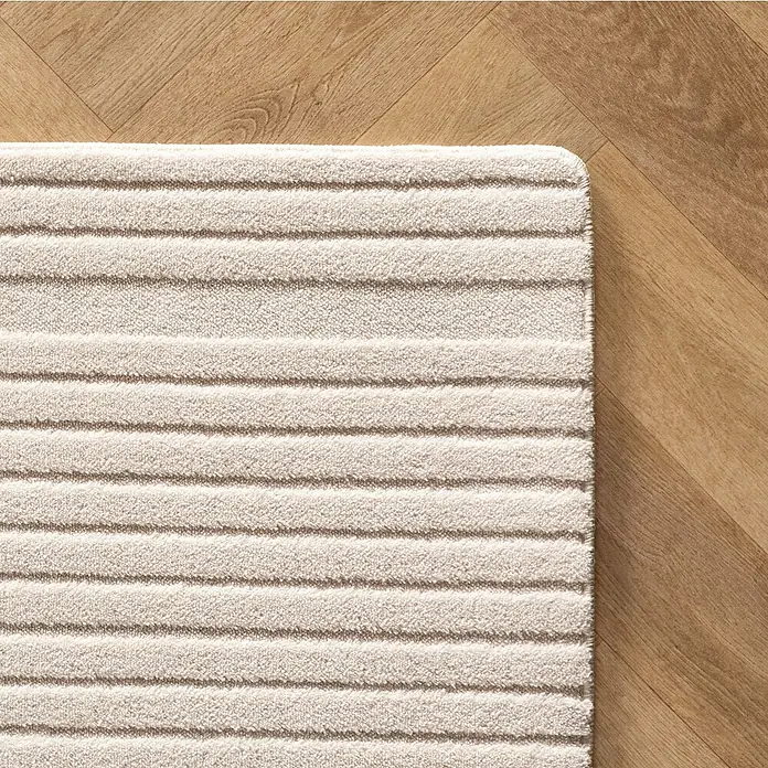 Kurzflorteppich Pompei 1612 - Creme, 340 x 240 cm, 100 % Polyester - Detailaufnahme eines beigen Badteppichs mit horizontalen Streifen auf einem hellen Holzboden. Der Teppich hat eine weiche, strukturierte Oberfläche.