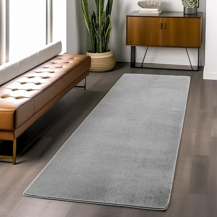 Kurzflorteppich Joy - Silber, 100 x 60 cm, 100 % Polyester - Ein grauer Teppich liegt in einem Wohnzimmer mit einem Ledersofa, einer Pflanze und einem Sideboard. Der Teppich verleiht dem Raum eine gemütliche Note.
