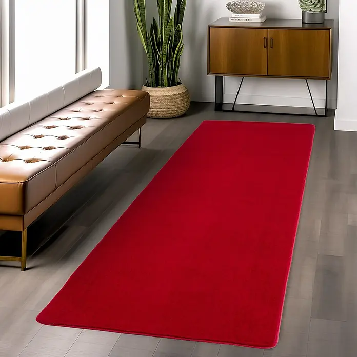 Kurzflorteppich Joy - Rot, 150 x 80 cm, 100 % Polyester - Roter Teppich liegt in einem stilvollen Wohnzimmer mit Ledersitzbank und Holzschrank.