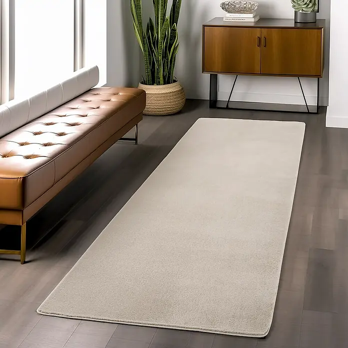 Kurzflorteppich Joy - Creme, 100 x 60 cm, 100 % Polyester - Ein heller, rechteckiger Teppich liegt auf einem dunklen Holzboden, neben einer braunen Bank und einem Holzschrank mit Pflanze.