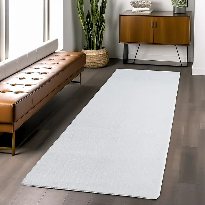 Kurzflorteppich Joy - Weiß, 100 x 60 cm, 100 % Polyester - Ein langer, weißer Teppich liegt auf einem dunklen Holzboden in einem modern eingerichteten Raum. Der Teppich ist der zentrale Fokus des Bildes.