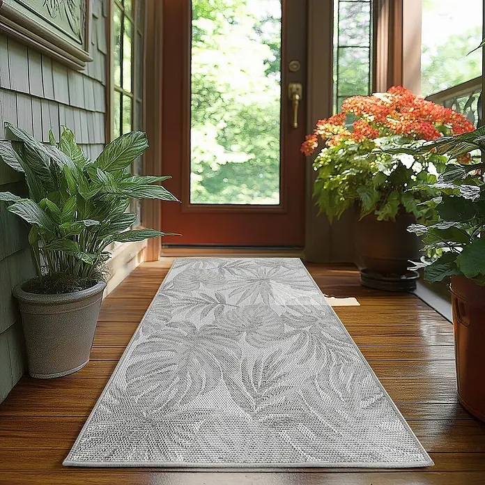 Outdoor-Teppich Desert 1302 - Grau, 150 x 80 cm, 100 % Polypropylen - Ein grauer Teppich mit Blattmuster liegt auf einem Holzboden in der Nähe einer Tür und Pflanzen, ein einladender Akzent für jeden Eingangsbereich.