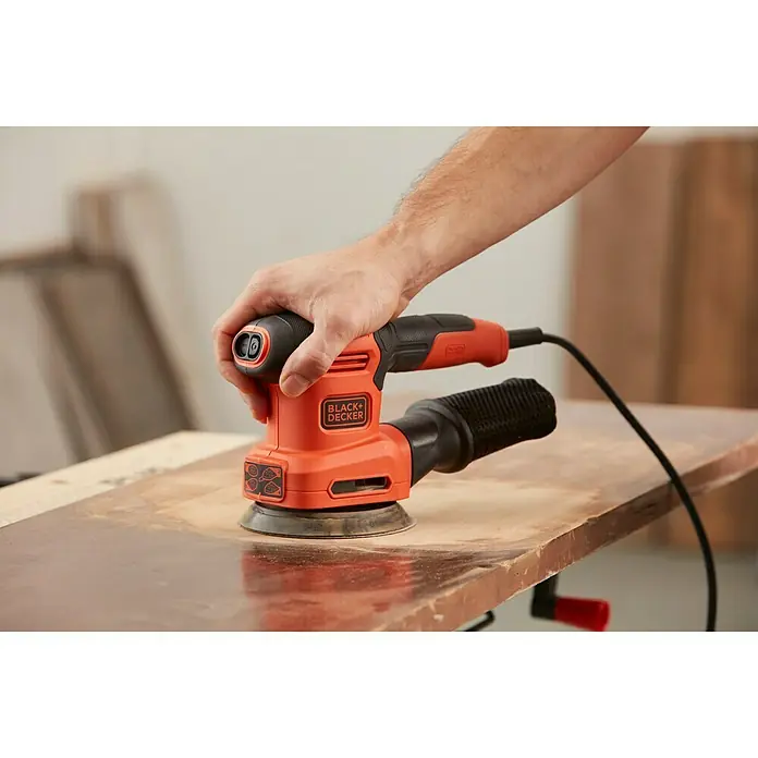 Black+Decker Lijadora multifunción eléctrica  BEW200-QS - 200 W, Número de oscilaciones marcha en vacío: 12.000 r.p.m., Diámetro circuito oscilante: 2 mm - Primer plano de una lijadora eléctrica Black+Decker en uso sobre una superficie de madera, sujetada por una mano.