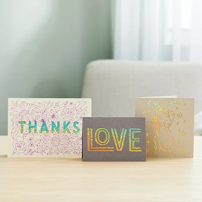Cricut Schneidematte Card Mat - L x B: 41 x 33 cm, Passend für: Cricut Maker, Cricut Explore Machines - Nahaufnahme von drei Grußkarten mit schimmernden Designs: "Thanks", "Love" und ein Hund mit Partyhut. Perfekt für besondere Anlässe.