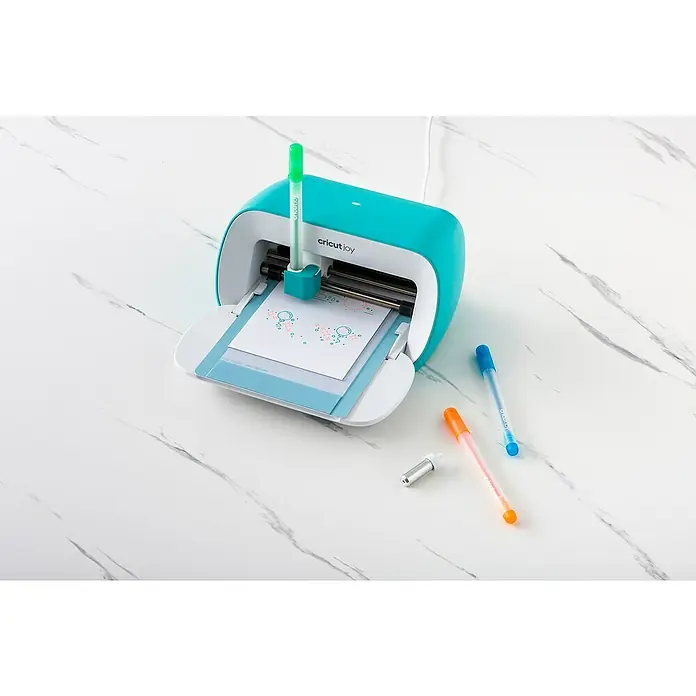 Cricut Gelroller-Set Glitzergelstifte - 10 -tlg., Mehrfarbig, 0,8 mm, Glitzernd - Cricut Joy Schneideplotter in Türkis mit Stiften und Schneidemesser. Der Plotter zeichnet Motive auf eine Karte, die auf einer Schneidematte liegt.