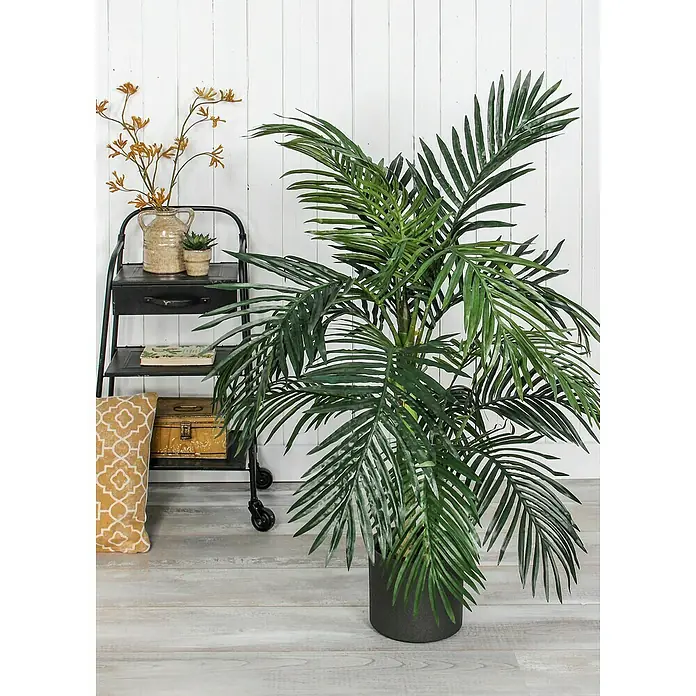 Kunstpflanze Areca Palme - Höhe: 150 cm, Grün, Kunststoff - Zimmerpflanze mit üppigen, grünen Blättern in einem dunklen Topf, neben einem Beistelltisch mit Dekorationen vor einer weißen Wand.