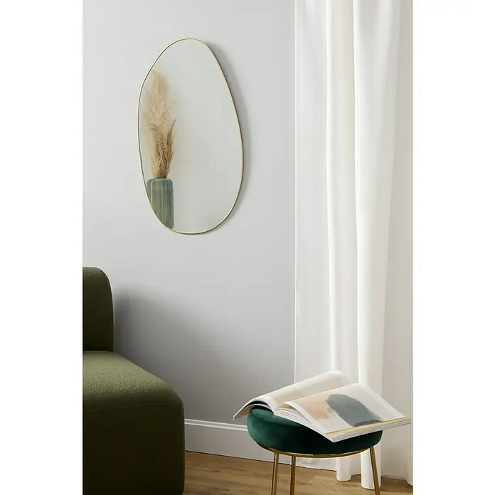 Espejo de pared Bode - 44 x 73 cm, Dorado - Espejo ovalado con marco dorado colgado sobre un sillón verde en una pared blanca, al lado de un taburete con revista.