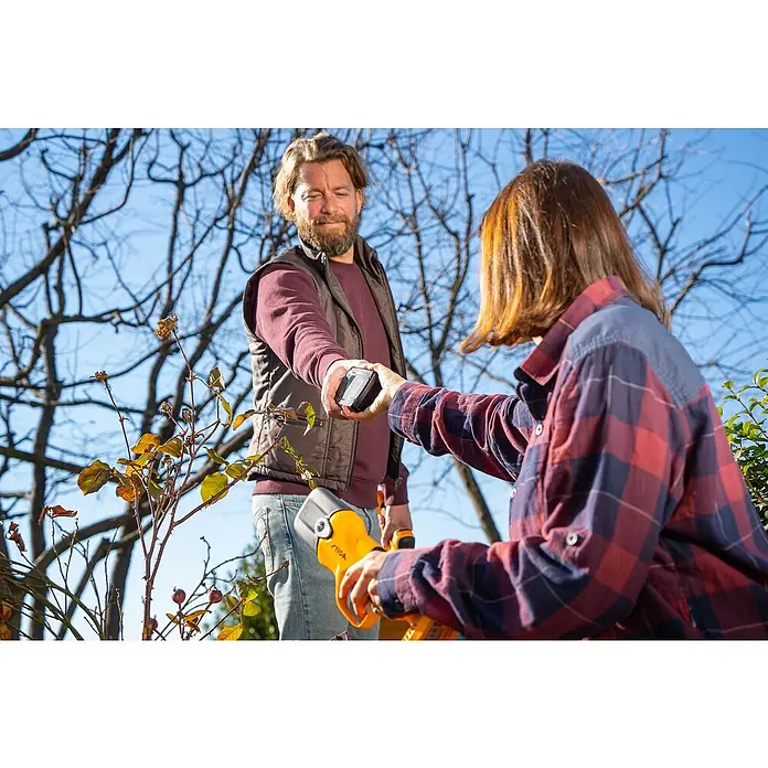 Stiga 100er Serie Akku-Astsäge PR100E & SC100E KIT -  - Ein Mann übergibt eine Batterie für eine STIGA-Heckenschere an eine Frau, beide sind im Garten mit Baumschnitt beschäftigt.