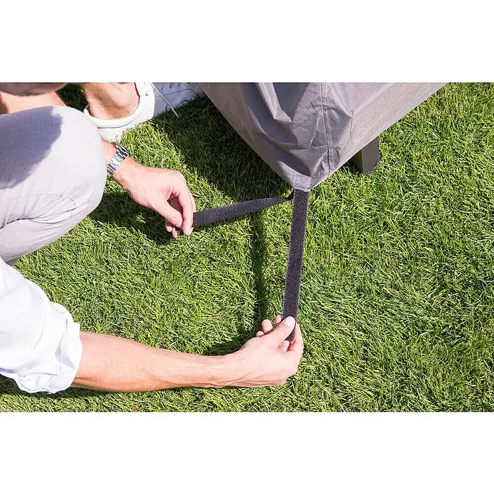 Barths Loungemöbel Schutzhülle - L x B x H: 215 x 215 x 70 cm, Polypropylen, Hellgrau - Detailaufnahme einer Person, die einen schwarzen Klettverschluss an einer grauen Gartenmöbelabdeckung auf grünem Gras befestigt.