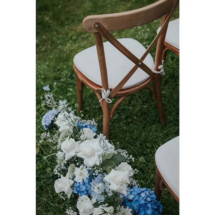 Stapelstuhl Crossback - 1 Stk., Ulmenholz, Braun, B x T x H: 48 x 47 x 89 cm - Eine Nahaufnahme von dekorierten Holzstühlen mit weißen Sitzkissen und einem Arrangement aus weißen Rosen und blauen Hortensien auf einer grünen Rasenfläche.