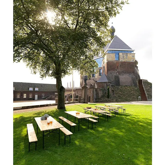 Feros Bierbank - Braun - Die Bierzelttische mit Bänken sind aus hellem Holz gefertigt und stehen auf einem grünen Rasen vor einem historischen Gebäude. Ideal für Feste!