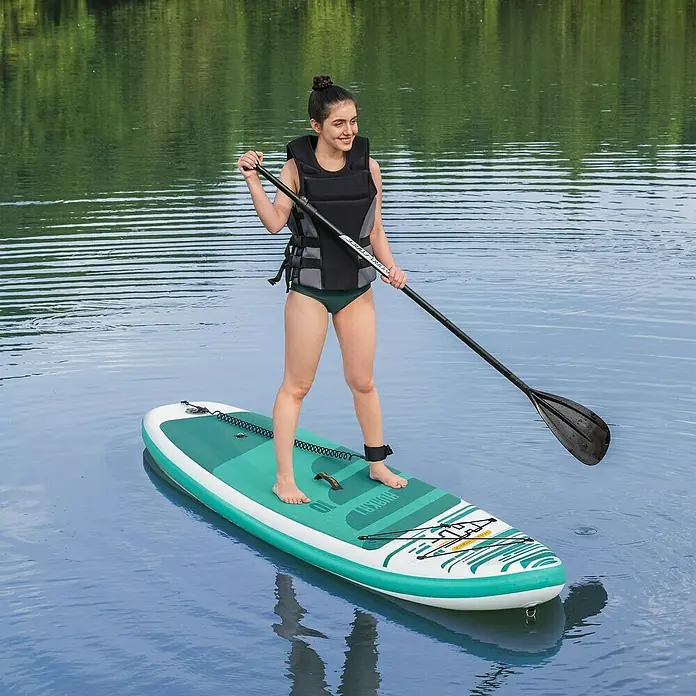 Bestway Hydro Force SUP-Board HuaKa'i - L x B x H: 305 x 84 x 15 cm, Nutzlast: 130 kg, Aufblasbar - Frau auf einem aufblasbaren Stand-Up-Paddleboard, das mit einem Paddel auf dem Wasser gleitet und eine Schwimmweste trägt.