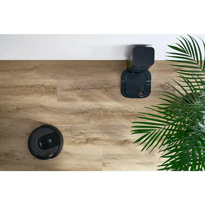 Vinylboden Silent - Sherman Eiche, Landhausdiele, 1.220 x 229 x 5,3 mm - Ein Roomba Saugroboter der Marke iRobot reinigt eine Holzbodenfläche mit einigen kleinen Schmutzpartikeln in der Nähe seiner Ladestation.