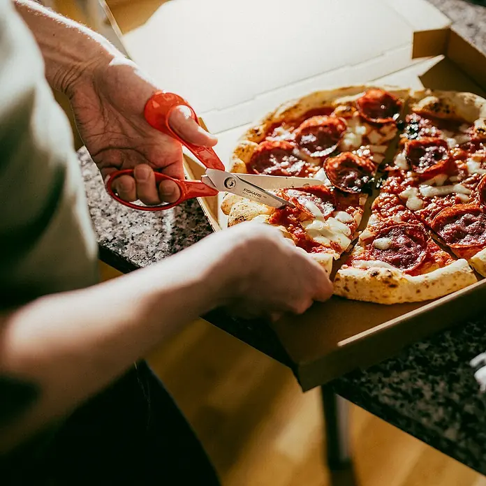 Fiskars Universalschere Classic - Länge: 21 cm, Rot - Eine Person schneidet eine Pizza mit Salami und Käse in einem Pizzakarton mit einer roten Schere.