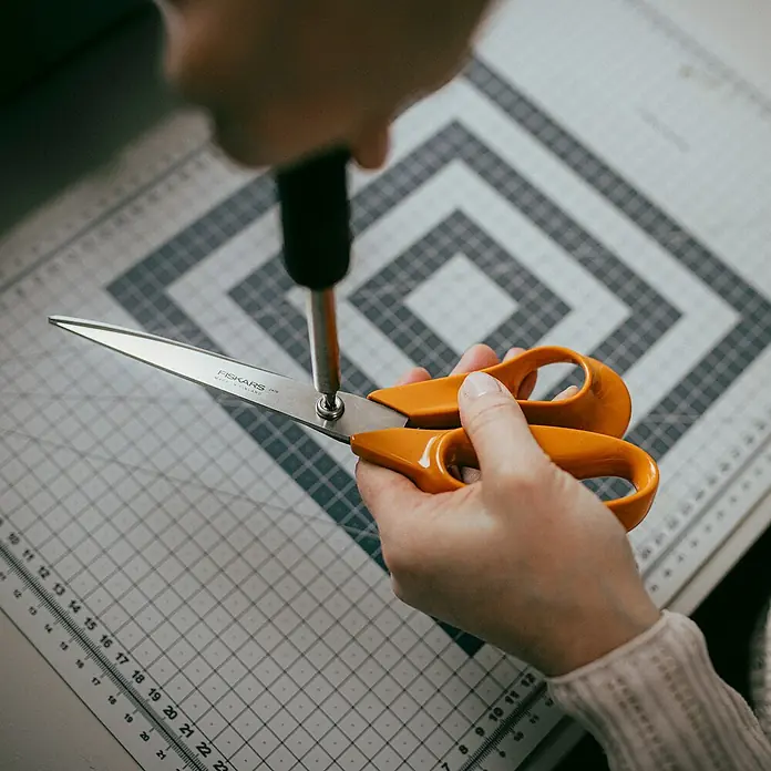 Fiskars Universalschere Classic - Rechtshänder, 25 cm, Farbe: Orange - Eine Nahaufnahme einer Person, die eine orangefarbene Fiskars-Schere mit einem Schraubenzieher auf einer Schneidematte repariert.