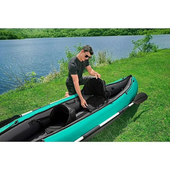 Bestway Hydro Force Kajak Ventura Elite™ X2 - L x B: 330 x 86 cm, Nutzlast: 200 kg, Geeignet für: 2 Personen - Ein Mann kniet neben einem türkis-schwarzen Kajak und platziert einen schwarzen Sitz darin, vor einem See und grüner Landschaft.