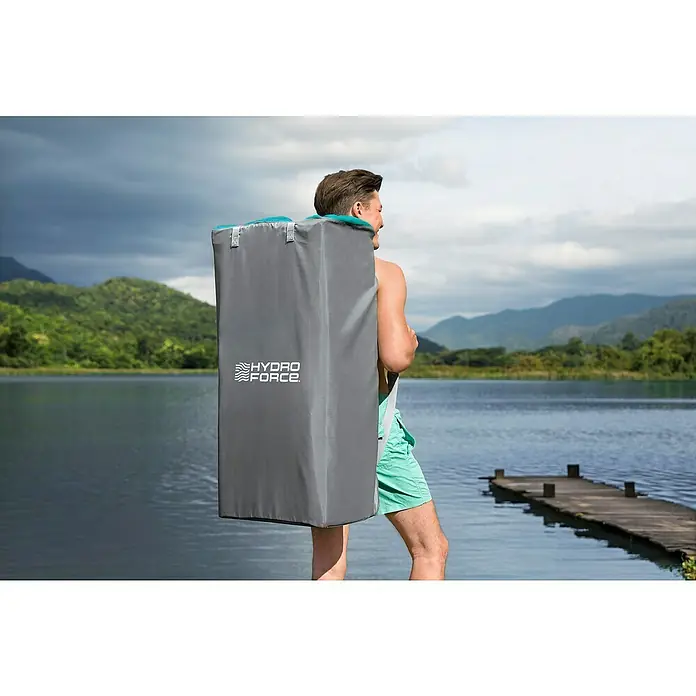 Bestway Hydro Force SUP-Board-Set Aqua Journey - L x B x H: 274 x 76 x 12 cm, Nutzlast: 100 kg, Aufblasbar, Blau/Weiß - Ein Mann trägt eine Hydro Force Tragetasche für ein aufblasbares Stand-Up-Paddle-Board vor einem See mit Bergen.