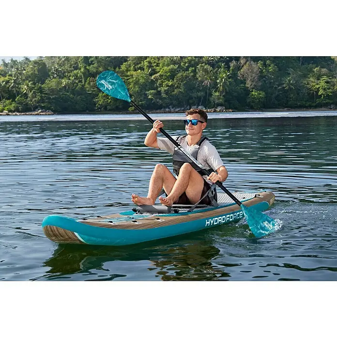 Bestway Hydro Force SUP-Board-Set Glider Elite™ mit Sitz - L x B x H: 381 x 79 x 15 cm, Nutzlast: 150 kg, Aufblasbar, Blau/Braun - Ein Mann paddelt auf einem Hydro-Force-Stand-Up-Paddleboard auf dem Wasser, umgeben von üppigem Grün.