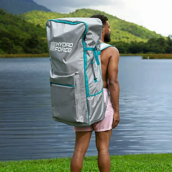 Bestway Hydro Force SUP-Board-Set Freesoul Elite™ mit Sitz - L x B x H: 335 x 84 x 15 cm, Nutzlast: 150 kg, Aufblasbar, Blau/Braun - Ein Mann mit einem übergroßen grauen Hydro Force Rucksack, der für Wassersportarten konzipiert ist, steht am Seeufer.