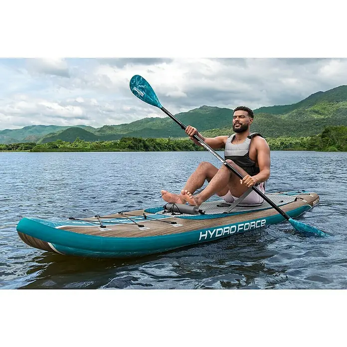 Bestway Hydro Force SUP-Board-Set Freesoul Elite™ mit Sitz - L x B x H: 335 x 84 x 15 cm, Nutzlast: 150 kg, Aufblasbar, Blau/Braun - Ein Mann paddelt auf einem Hydro Force Stand-Up-Paddleboard auf einem ruhigen See mit Bergen im Hintergrund.