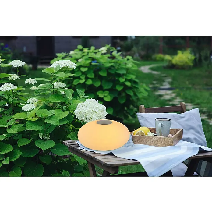 Globo LED-Tischleuchte Laela - 0,5 W, Weiß, RGBW, Ø x H: 20 x 11,7 cm - Diese Szene zeigt eine dekorative Gartenleuchte, die auf einem Tisch neben Getränken und weißen Hortensien sitzt, perfekt für stimmungsvolle Gartenabende.